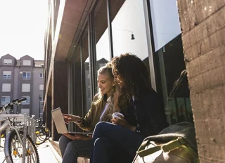 Deux femme assises dans la rue en train de sourire devant un ordinateur portable